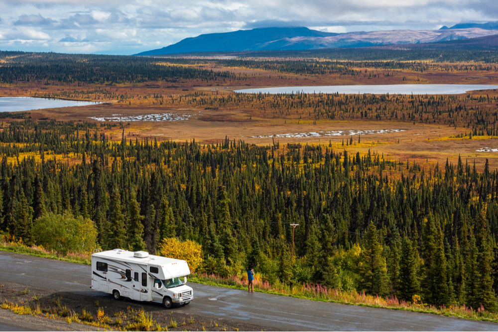 RV, motorhome, caravan parking next to a road in a parking lot in Alaska with spectacular, beautfiul background with lakes and conifer forest, blue sky and clouds