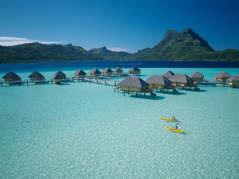 overwater bungalows in Bora Bora with green mountain in background and kayakers in foreground and very clear ocean