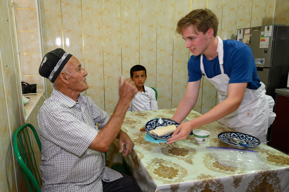 american white teenage boy serves food to older Uzbek man in a kitchen in Bukhara Uzbekistan during a shared cooking experience