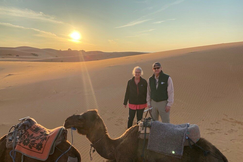 man and woman couple standing behind a camel in the desert at sunset in Morocco