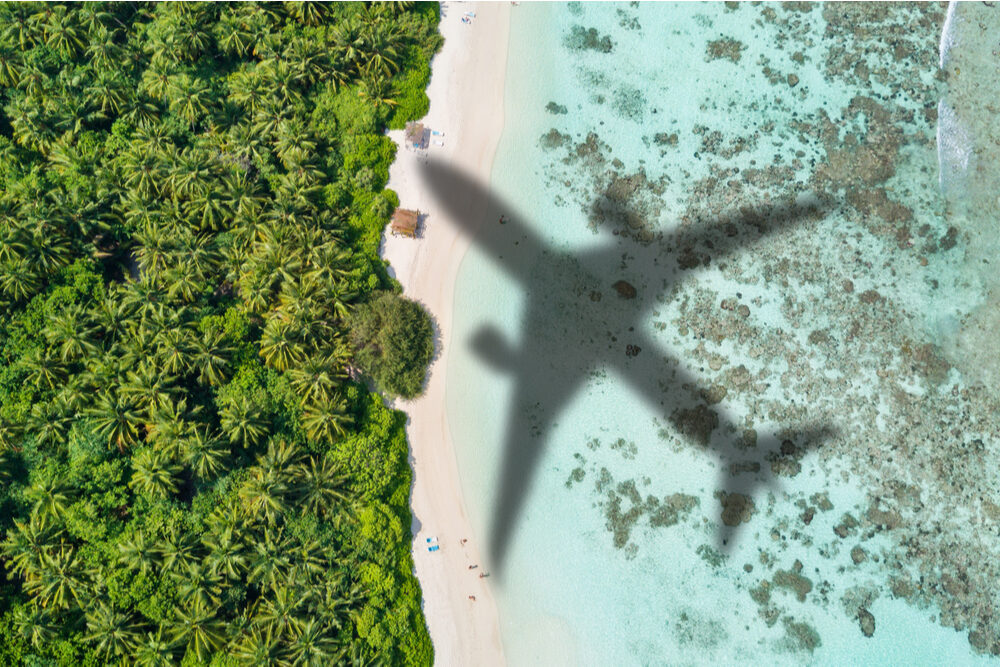 Shadow of an airliner over a blue water and a white-sand beach.