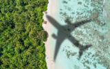 Shadow of an airliner over a blue water and a white-sand beach.