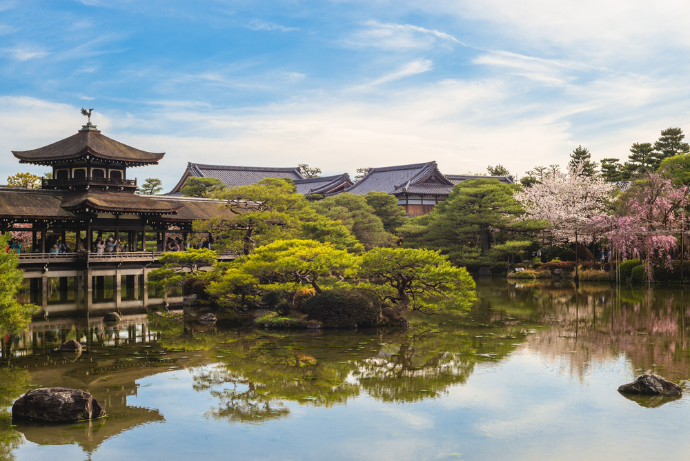 Japanese garden with trees, cherry blossoms, and buildings next to the river.