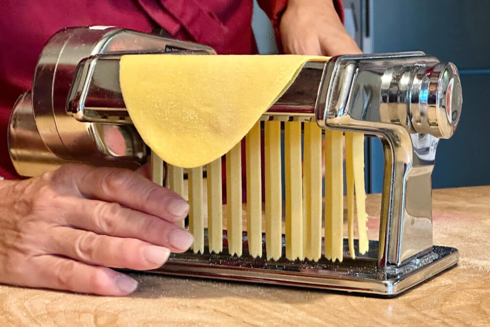 Fresh pasta dough being cut into strips with a hand-cranked pasta machine.