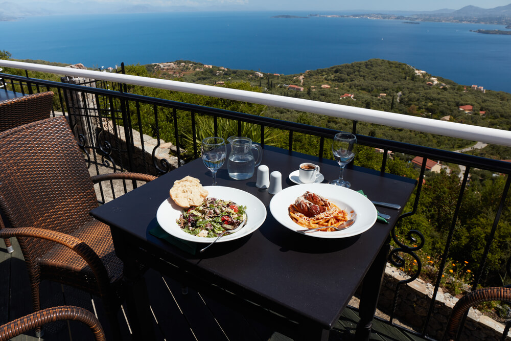 Outdoor restaurant table in Corfu overlooking the sea, set with a plate of pastitsada.